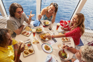 A group of Sailors eating together at The Galley, the onboard food hall. 5 adults are gathered around a round table with big windows behind them. The table is full of food items like burgers, tacos, ramen, and salads.